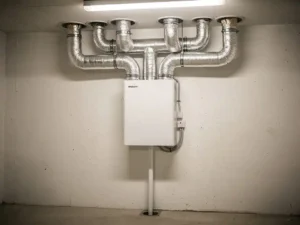 Wit MVHR-unit gemonteerd op muur in technische ruimte, met zilveren flexibele kanalen naar plafondventielen en condensaatafvoer.