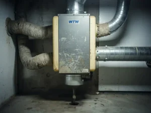 Verouderd WTW-ventilatie-unit in Nederlandse meterkast met vergeeld kunststof behuizing, verstopte geïsoleerde kanalen en condensafvoer.