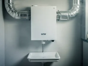 WTW warmteterugwinunit aan muur in technische ruimte, met zilveren geïsoleerde kanalen en condensaatafvoer in opvangbak.