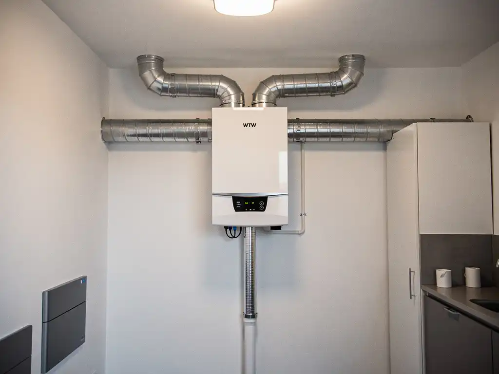 WTW-unit gemonteerd op muur in technische ruimte van modern appartement, met zilveren geïsoleerde kanalen en condensafvoer.