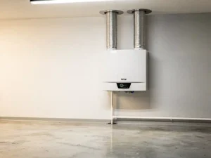 Witte WTW-unit aan muurwand in technische ruimte, verbonden met zilveren flexibele luchtkanalen naar plafondopeningen en condensaatafvoer langs de plint.
