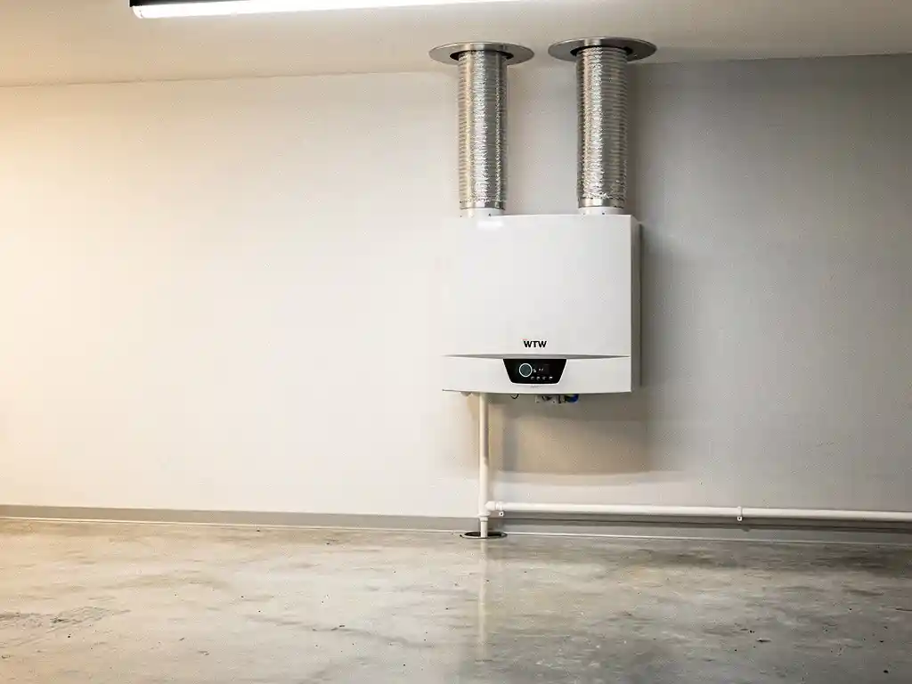 Witte WTW-unit aan muurwand in technische ruimte, verbonden met zilveren flexibele luchtkanalen naar plafondopeningen en condensaatafvoer langs de plint.