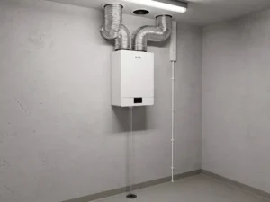 WTW warmteterugwinunit op muur in technische ruimte, aangesloten met zilveren flexibele luchtkanalen en condensaatafvoer.