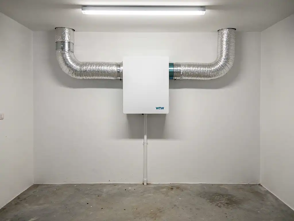 Witte WTW-unit aan muurbevestigd in technische ruimte met zilveren geïsoleerde kanalen en condensafvoerbuis.