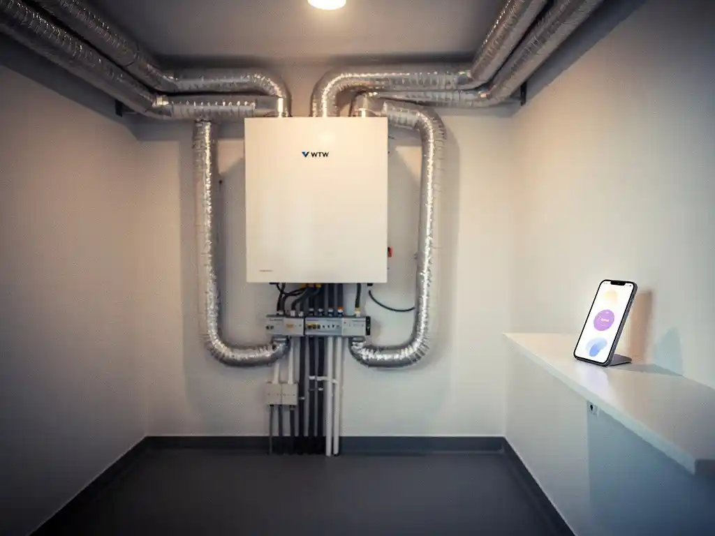 WTW warmteterugwinunit op muur in technische ruimte, met zilveren isolatiebuizen en smartphone met ventilatiebesturing op plank.